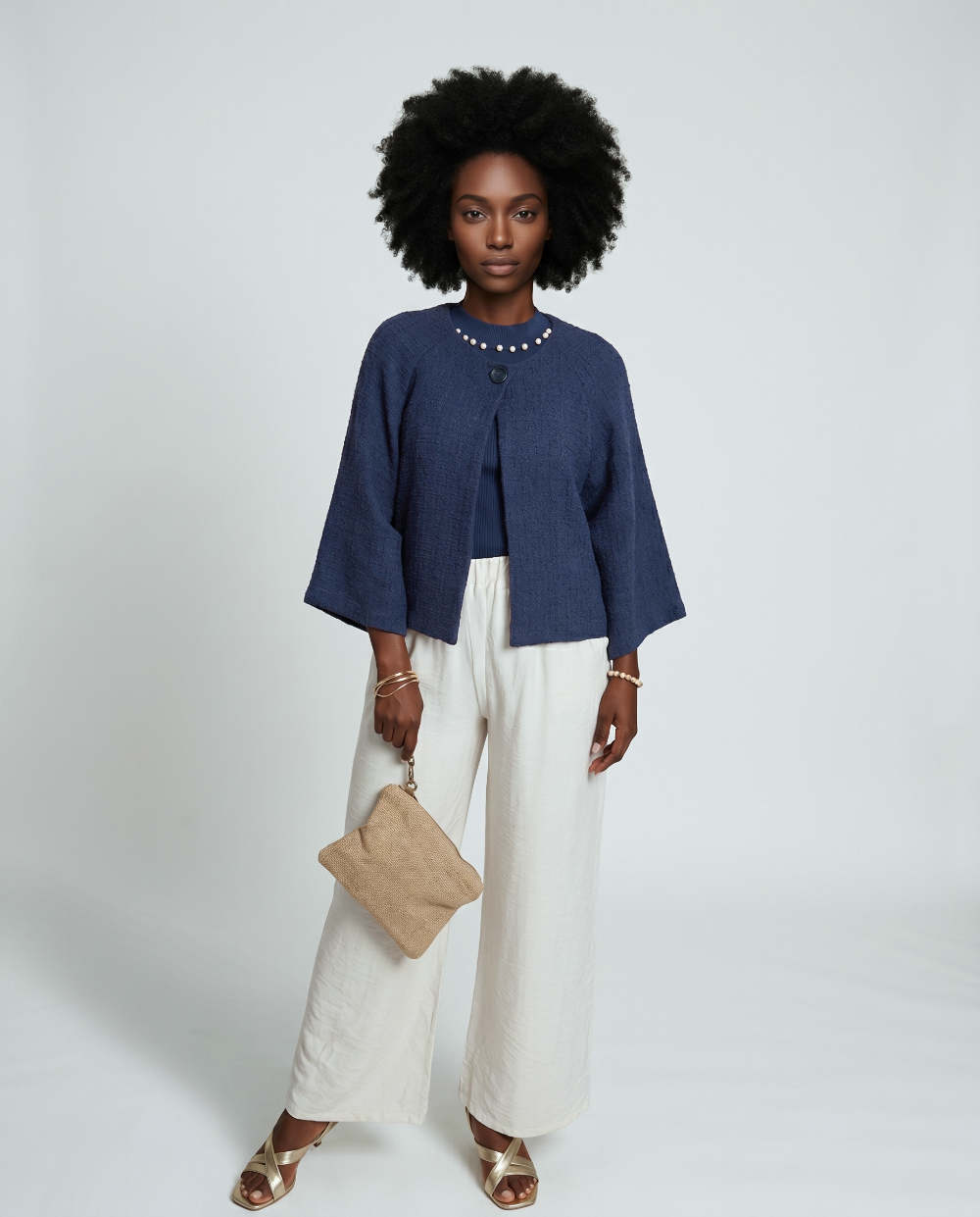 Woman wearing a blue top and white pants with a beige clutch against a white background